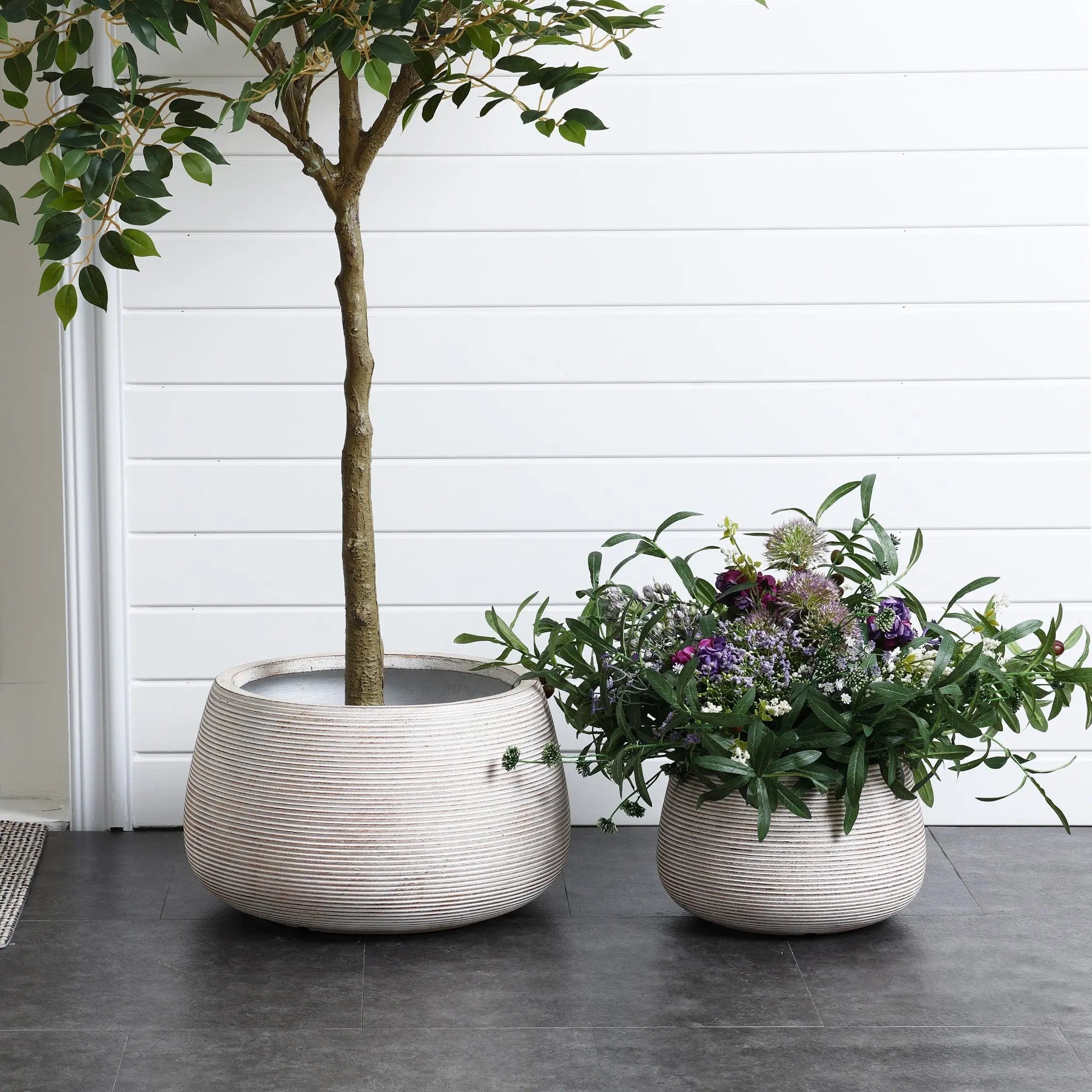 Two round, textured planters rest on a dark floor; the left planter cradles a small tree, while the right showcases various flowering plants. A sleek white panel wall provides a minimalist backdrop.
