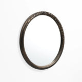 A LuxenHome Aged Wood Finish Crater Pattern Round Wall Mirror hangs on a white wall. The dark brown frames raised pattern gives it an artistic, industrial-inspired look.