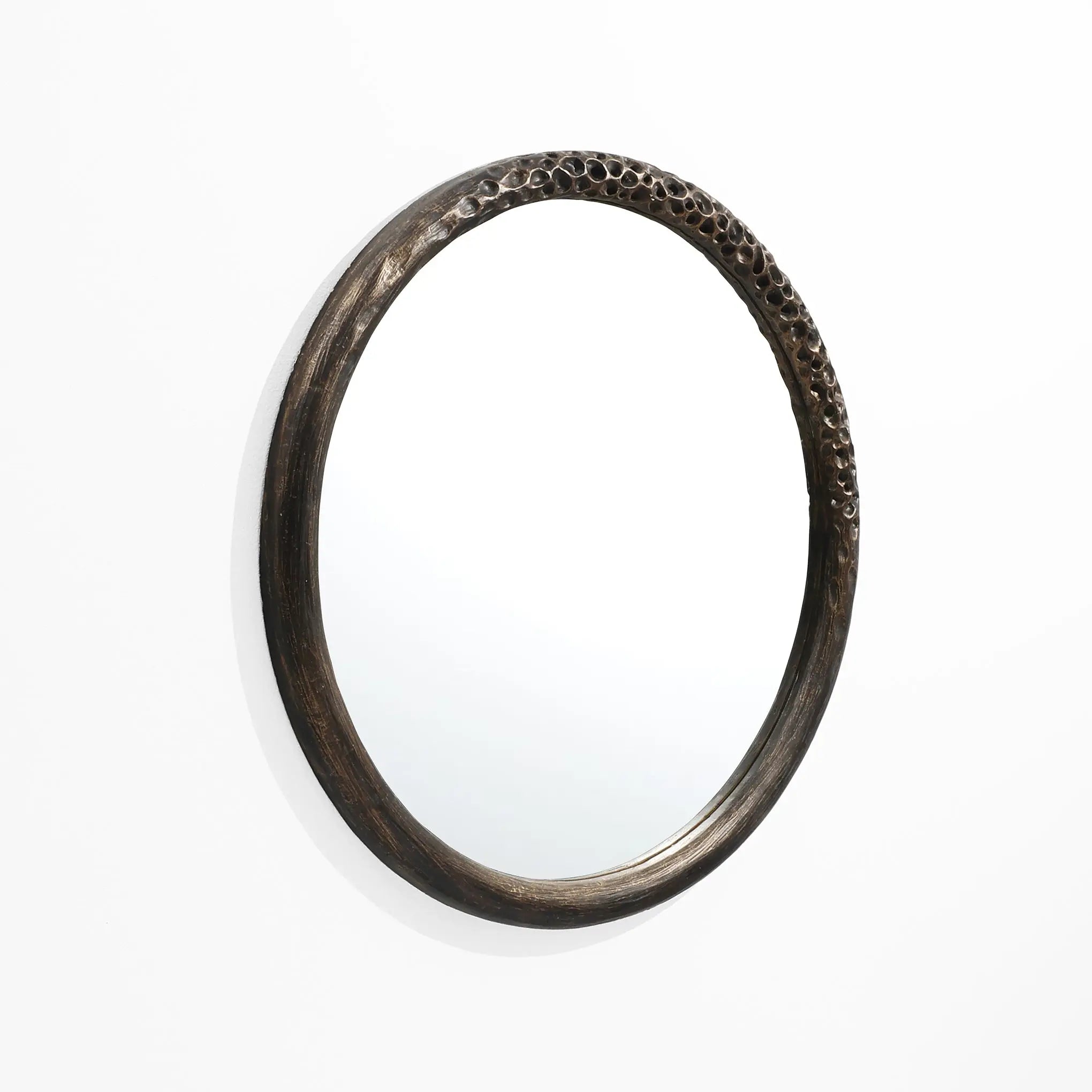 A LuxenHome Aged Wood Finish Crater Pattern Round Wall Mirror hangs on a white wall. The dark brown frames raised pattern gives it an artistic, industrial-inspired look.