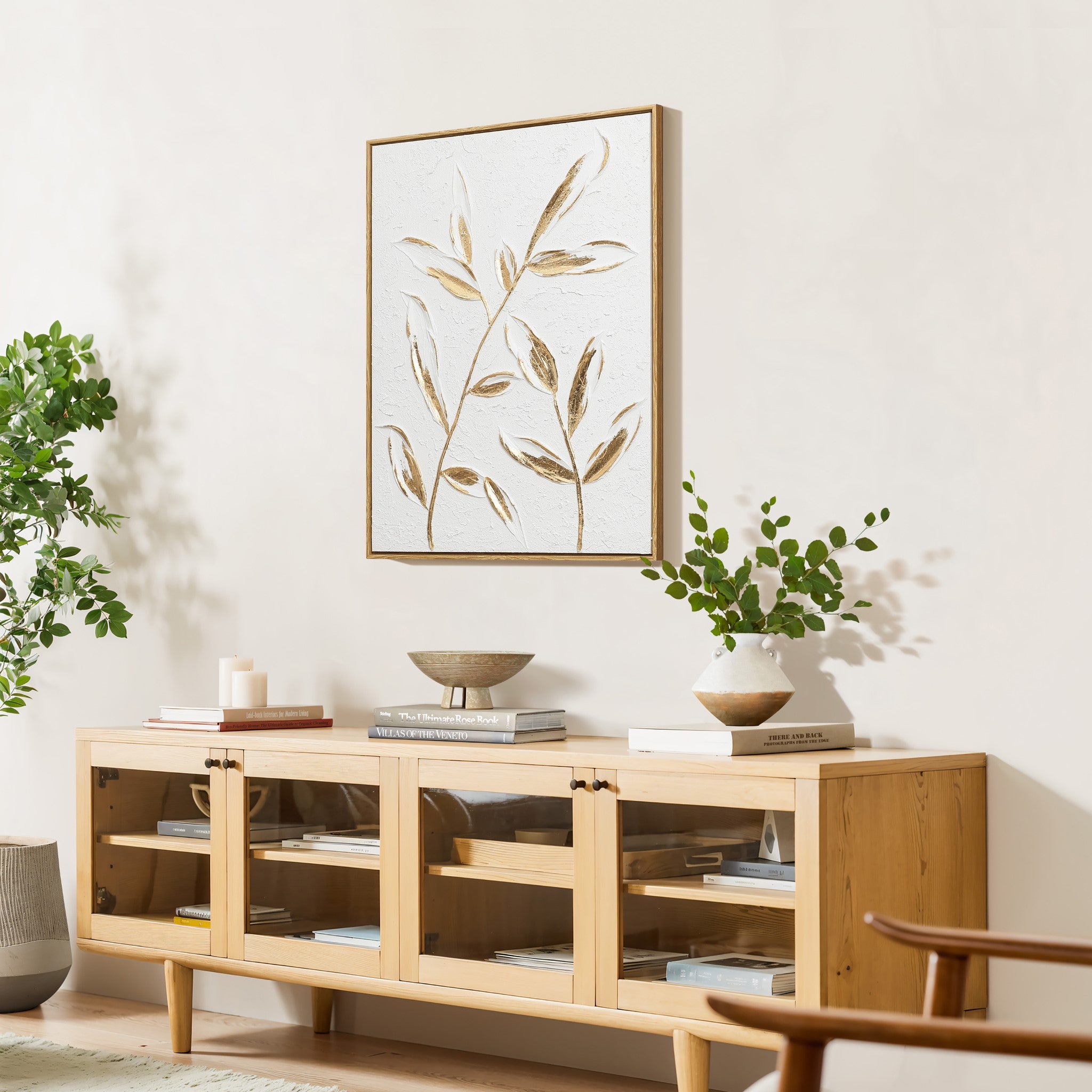 Wooden sideboard with glass doors, topped with decorative items. A potted plant and candle are on the left, with a painting of leaves featuring a gold leaf motif above. Greenery is on either side, creating a harmonious blend of nature and elegance.