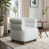 The LuxenHome Tia ivory barrel accent chair with wooden ball legs sits on a textured rug in a sunlit room. Large windows illuminate a side table featuring a mug, potted plants, and framed art on the wall.