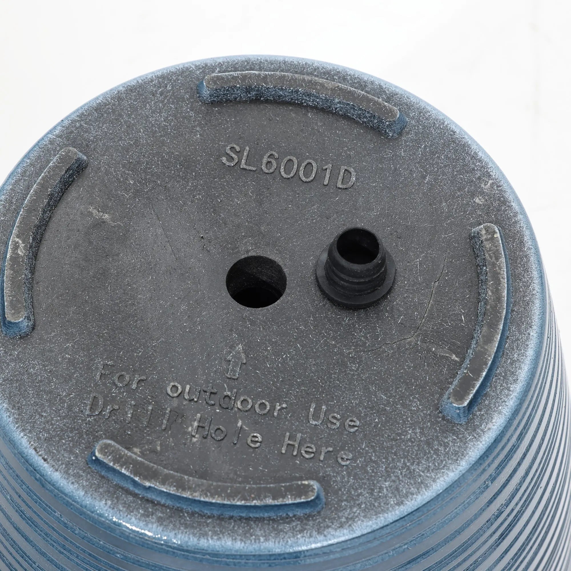 Close-up of the bottom of a blue plastic plant pot, featuring a drainage hole and the text For outdoor use Dr 1 Hole Here alongside the model number SL6001D. This decorative planter boasts a UV-protected finish, making it perfect for both indoor and outdoor use.