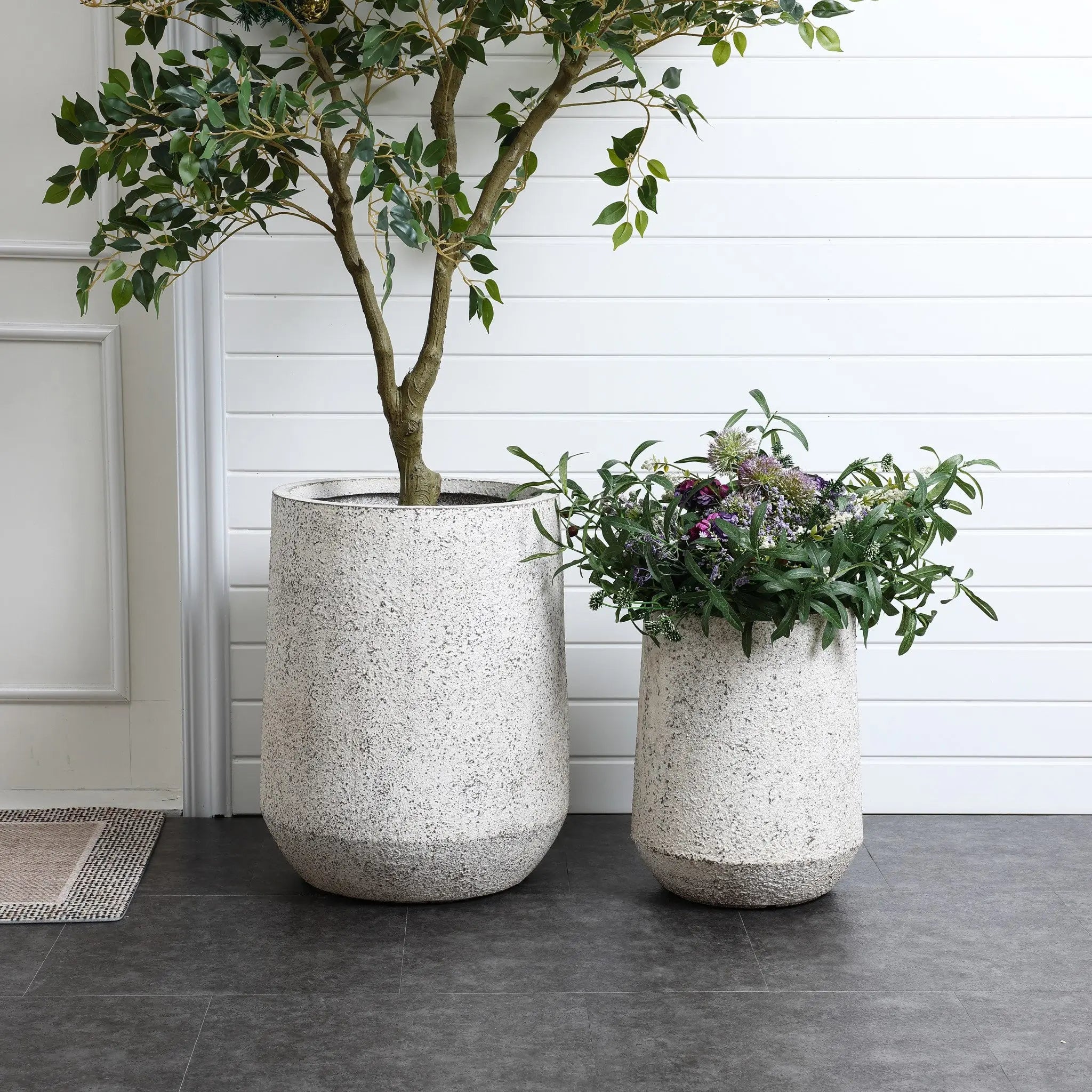 Two large, textured MgO planters on a dark floor; one houses a leafy tree, the other holds a plant with green leaves and small purple flowers. Their modern design features a gray and white speckled texture. A white paneled wall serves as the backdrop.