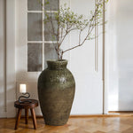 A large stoneware vase with branches complements the rustic charm of a small wooden table and lamp. They rest on a light wood herringbone floor, set against a white paneled wall and glass window.