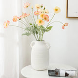 A white ceramic vase exuding rustic charm holds pink and yellow flowers on a round white table, next to a rolled magazine.