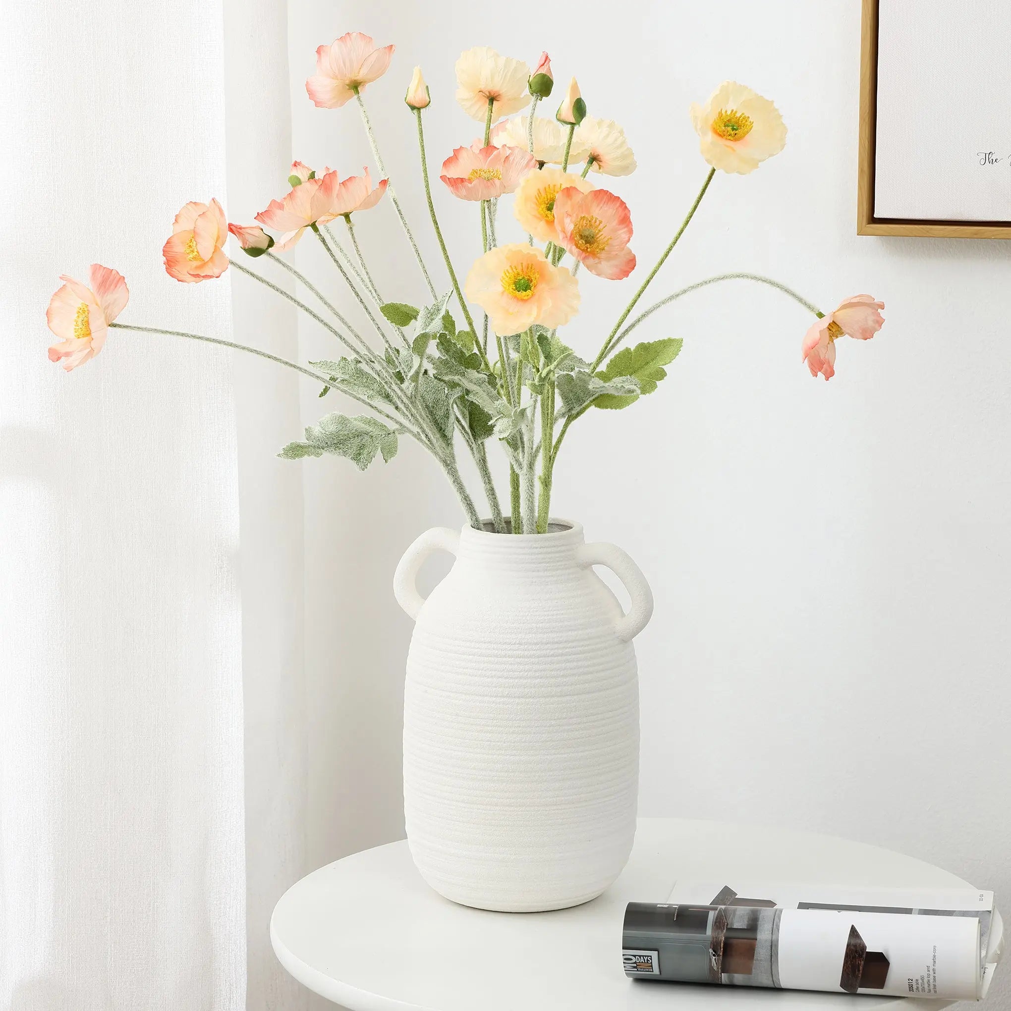 A white ceramic vase exuding rustic charm holds pink and yellow flowers on a round white table, next to a rolled magazine.
