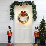 A white door adorned with an LED-lit holiday wreath and garland. Two nutcracker figures flank the door, while a small Christmas tree brightens the scene of this charming outdoor holiday decoration.