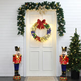 A white door adorned with an LED-lit holiday wreath and garland. Two nutcracker figures flank the door, while a small Christmas tree brightens the scene of this charming outdoor holiday decoration.