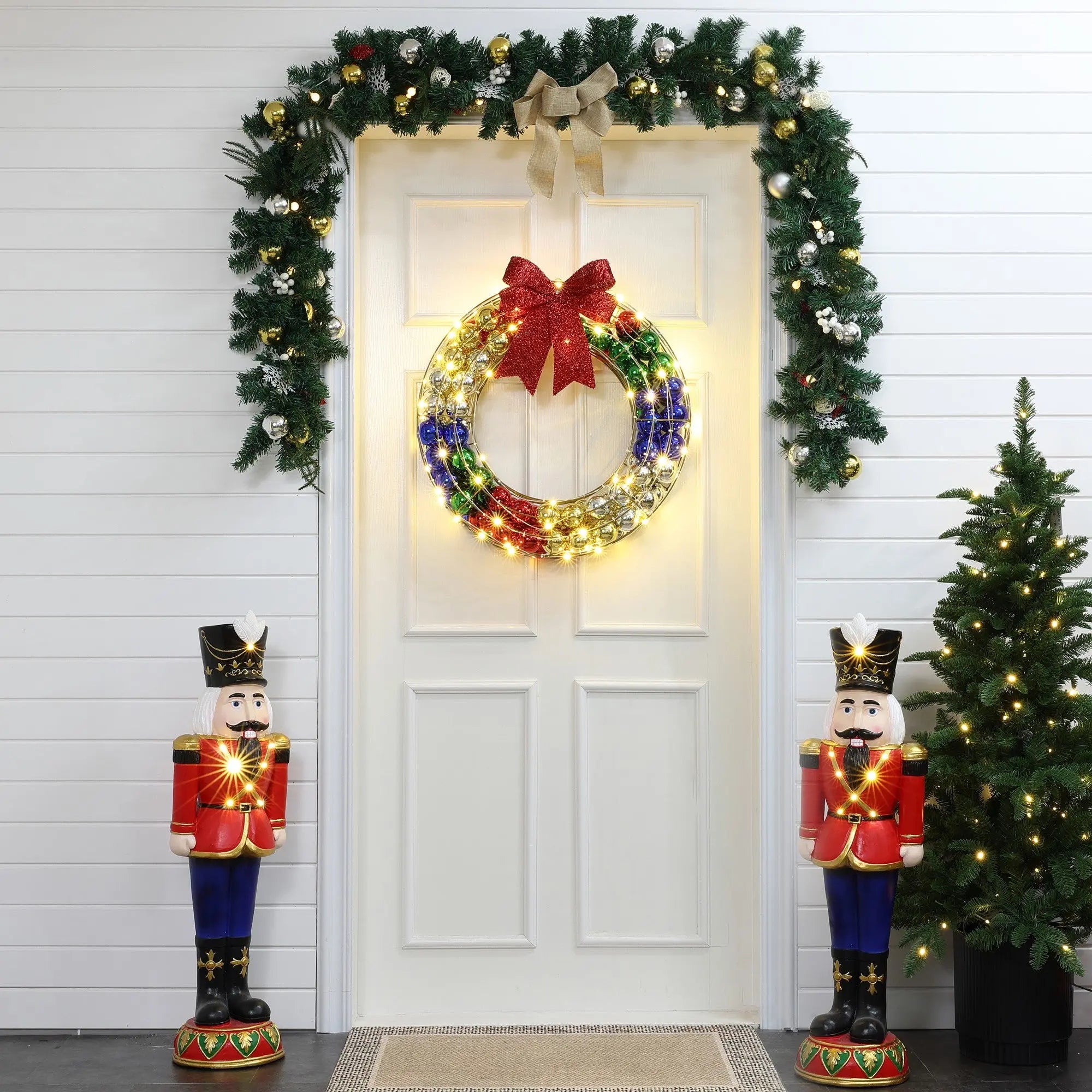 A white door adorned with an LED-lit holiday wreath and garland. Two nutcracker figures flank the door, while a small Christmas tree brightens the scene of this charming outdoor holiday decoration.