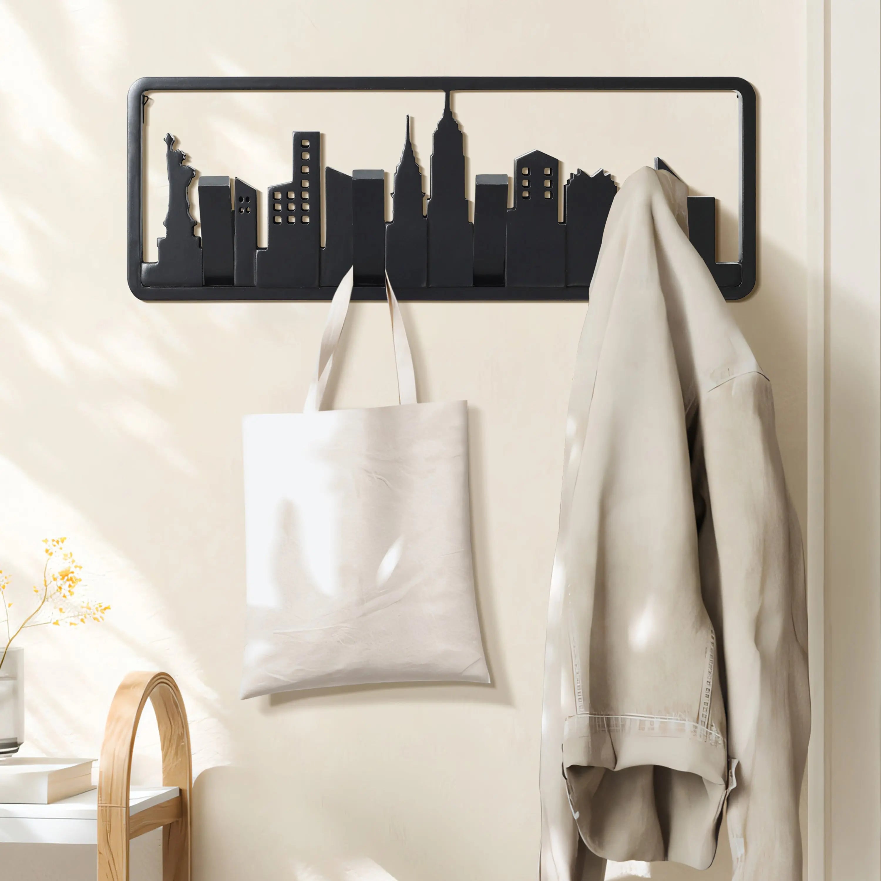 A black silhouette of a city skyline wall hook crafted from solid fir wood, with flip-down hooks holding a white tote bag and beige jacket. It is mounted on a light-colored wall with a small plant nearby.