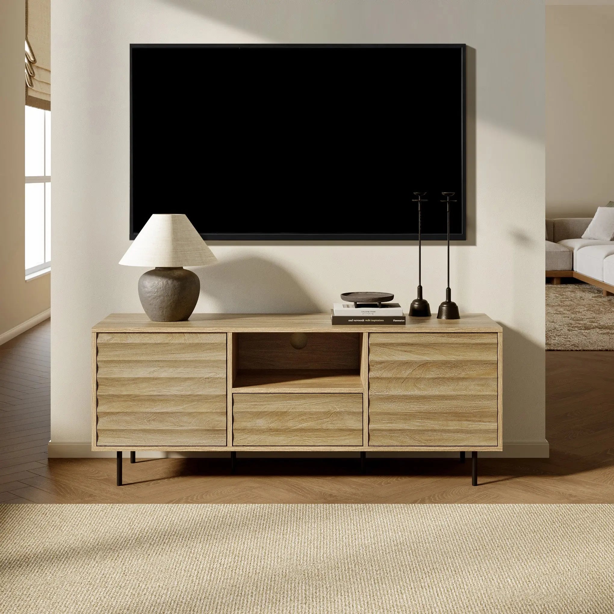 A modern living room showcases a large flat-screen TV mounted above a wooden console crafted from light walnut-colored wood. The console is adorned with a lamp, books, and candle holders, and even includes a cable management hole for tidy organization.