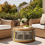 An idyllic seating area features two wicker chairs and a round, acacia wood table adorned with a vase of white flowers and a bowl of green apples. The hand-woven rope details complement the lush greenery and houses visible in the background, merging indoor and outdoor serenity.