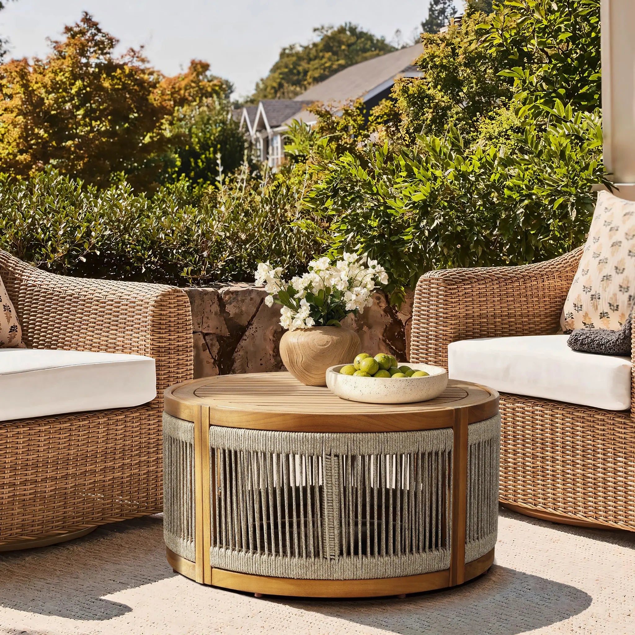 An idyllic seating area features two wicker chairs and a round, acacia wood table adorned with a vase of white flowers and a bowl of green apples. The hand-woven rope details complement the lush greenery and houses visible in the background, merging indoor and outdoor serenity.