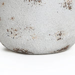 A close-up of the base of a round planter reveals its weathered, textured white surface, highlighted by patches of peeling paint and spots of rust. Its vintage minimalist style is beautifully accentuated, set against a plain white background.