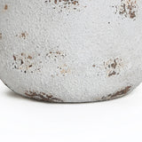 A close-up of the base of a round planter reveals its weathered, textured white surface, highlighted by patches of peeling paint and spots of rust. Its vintage minimalist style is beautifully accentuated, set against a plain white background.