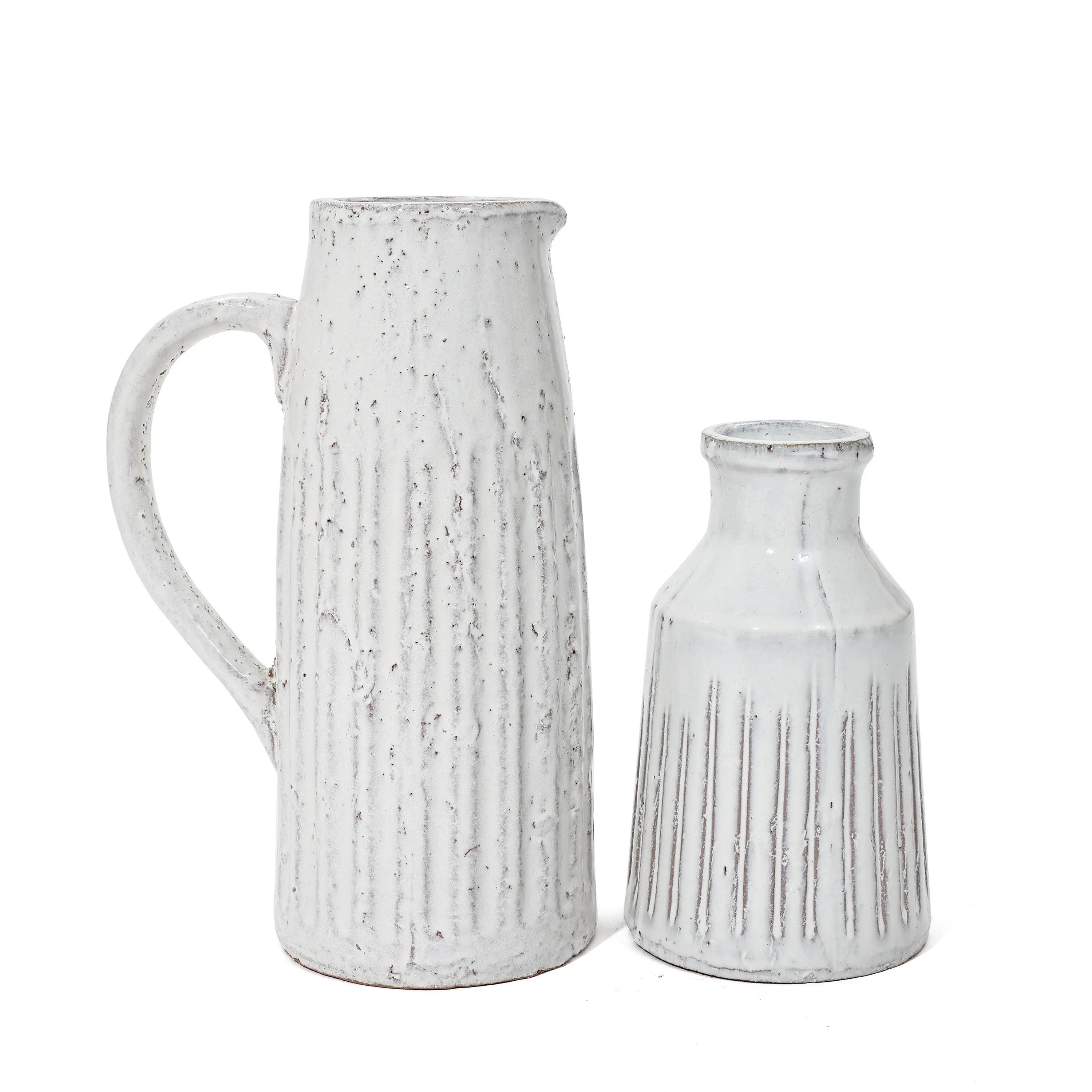 Two white ceramic vases with vertical ridges exude rustic charm. The taller vase, reminiscent of a milk jug with its handle and spout, and the smaller one, featuring a round opening, both boast a textured, slightly distressed finish that adds to their vintage design.