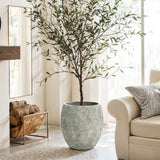 A tall potted tree in the LuxenHome Rustic Multi-Color Cement 15.4-Inch Round Planter stands by a white couch in a bright living room. A woven basket with logs sits on the floor, and large windows let abundant natural light flood in.