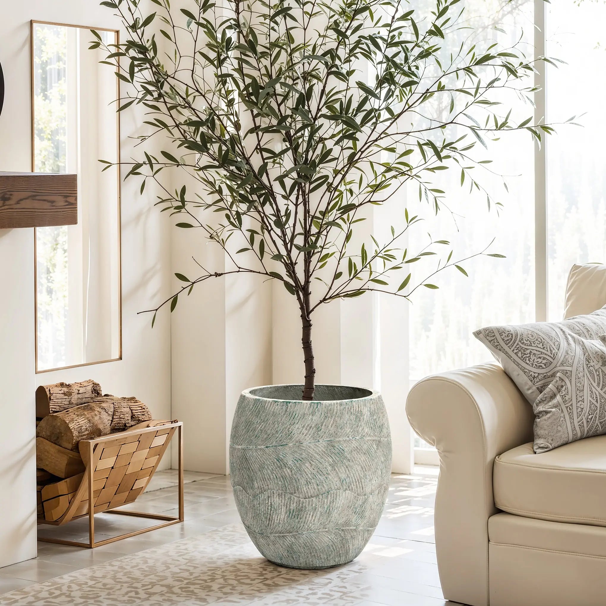 A tall potted tree in the LuxenHome Rustic Multi-Color Cement 15.4-Inch Round Planter stands by a white couch in a bright living room. A woven basket with logs sits on the floor, and large windows let abundant natural light flood in.