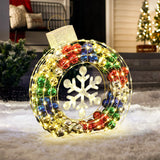 A large, illuminated ornament-shaped holiday decoration with multicolored lights and a central snowflake design sits on a snowy porch. Outdoor lights add sparkle alongside a Christmas tree that glows warmly in the background.