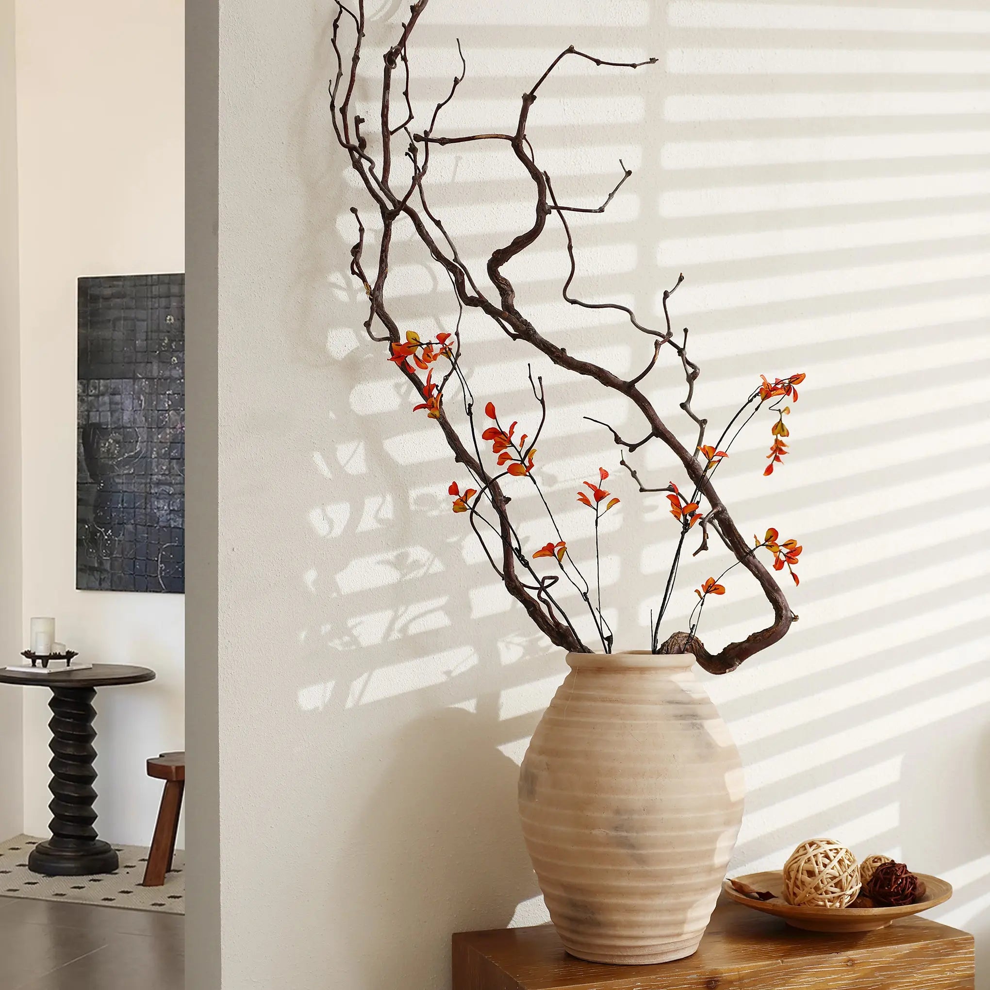 A LuxenHome rustic beige ribbed terracotta urn vase, 13.2 h, with long branches and small orange flowers sits on a wooden table by a white wall. Sunlight from a nearby window highlights its texture with striped shadows. A small round table and abstract wall art are in the background.