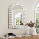 A decorated vase with dried flowers and a pair of sunglasses sit on a marble-topped wooden cabinet below an arched rectangular wall mirror, reflecting a window with greenery outside—perfect for stylish home decor.