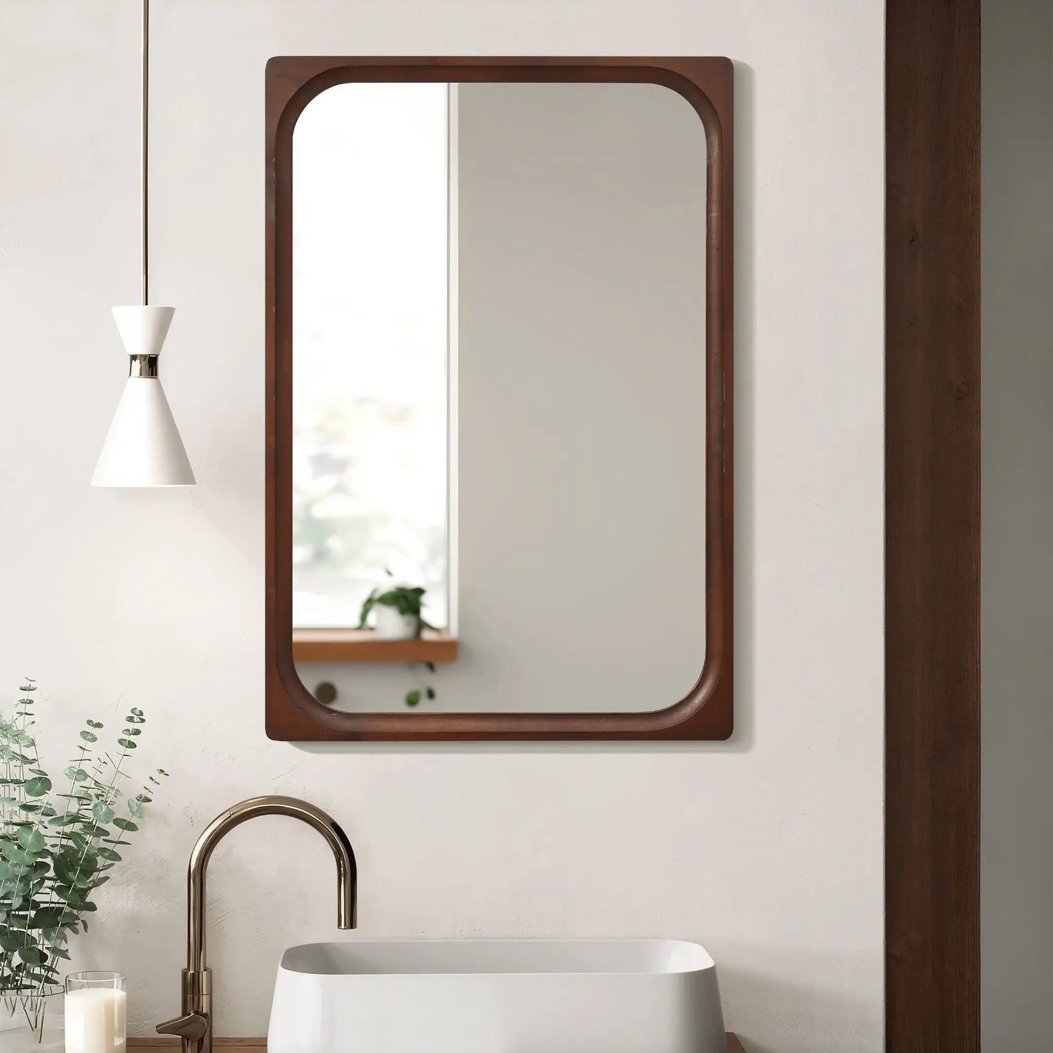 A rectangular wall mirror with a rubberwood frame is mounted on a light-colored wall above a white sink. A single hanging lamp and a potted plant are thoughtfully positioned to the left, adding charm to this elegant piece of home decor.