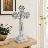 A decorative standing cross with intricate carvings is placed on a wooden table, embodying the essence of the Christian faith, next to a potted plant. A framed angel painting hangs on the light-colored wall in the background.