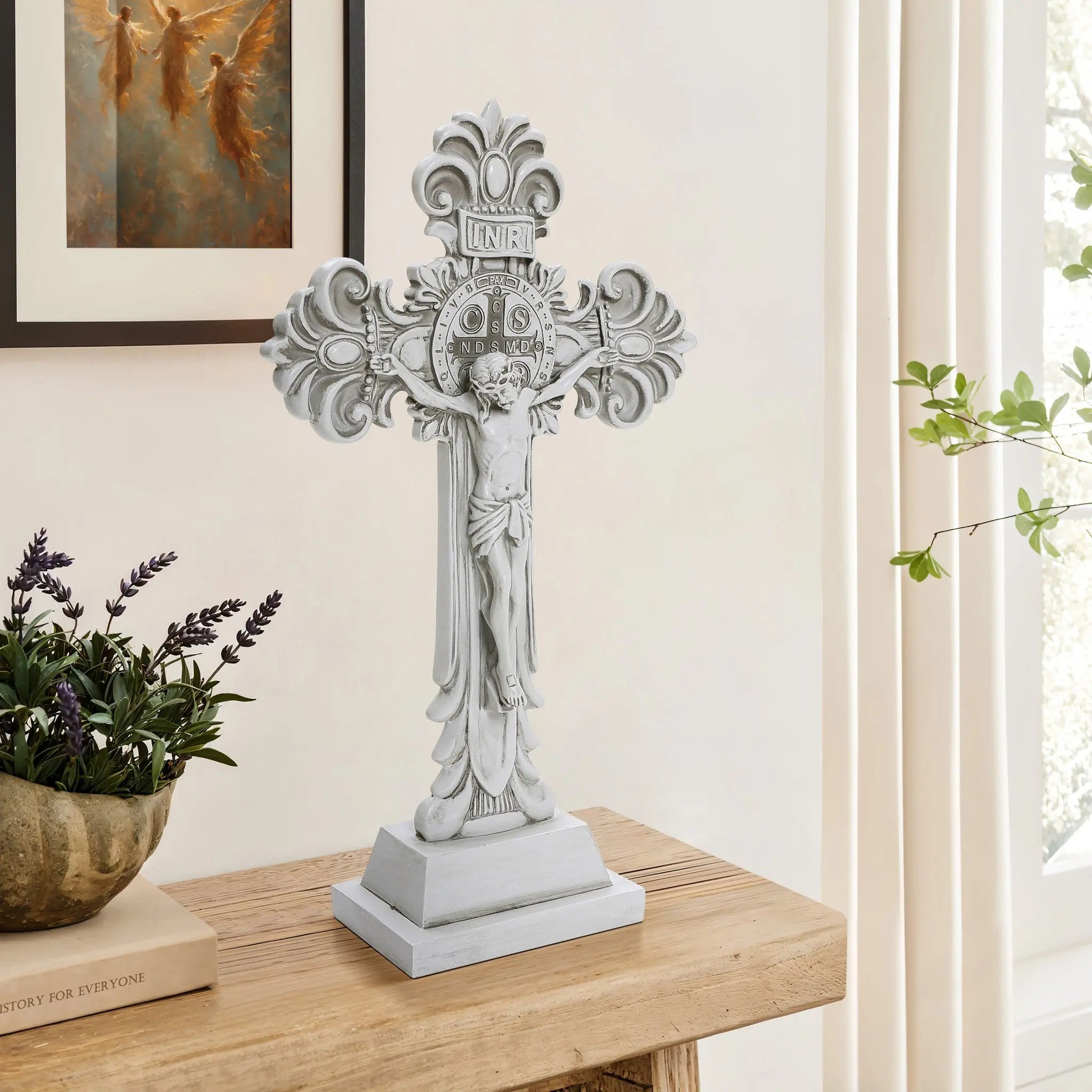 A decorative standing cross with intricate carvings is placed on a wooden table, embodying the essence of the Christian faith, next to a potted plant. A framed angel painting hangs on the light-colored wall in the background.