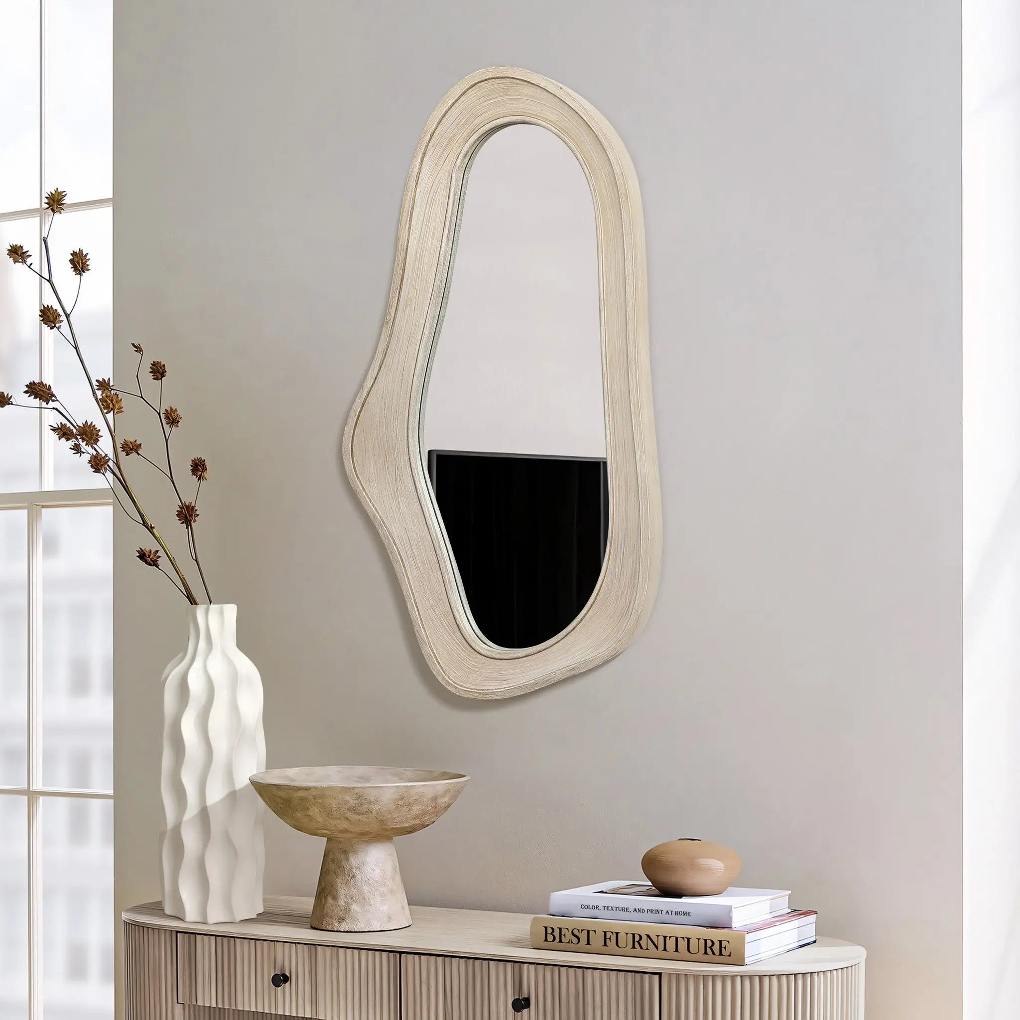 An asymmetrical mirror with an oval, irregular shape and a natural finish wood frame hangs above a wooden console. A white vase, decorative bowl, and a small round object complete this modern home decor arrangement.
