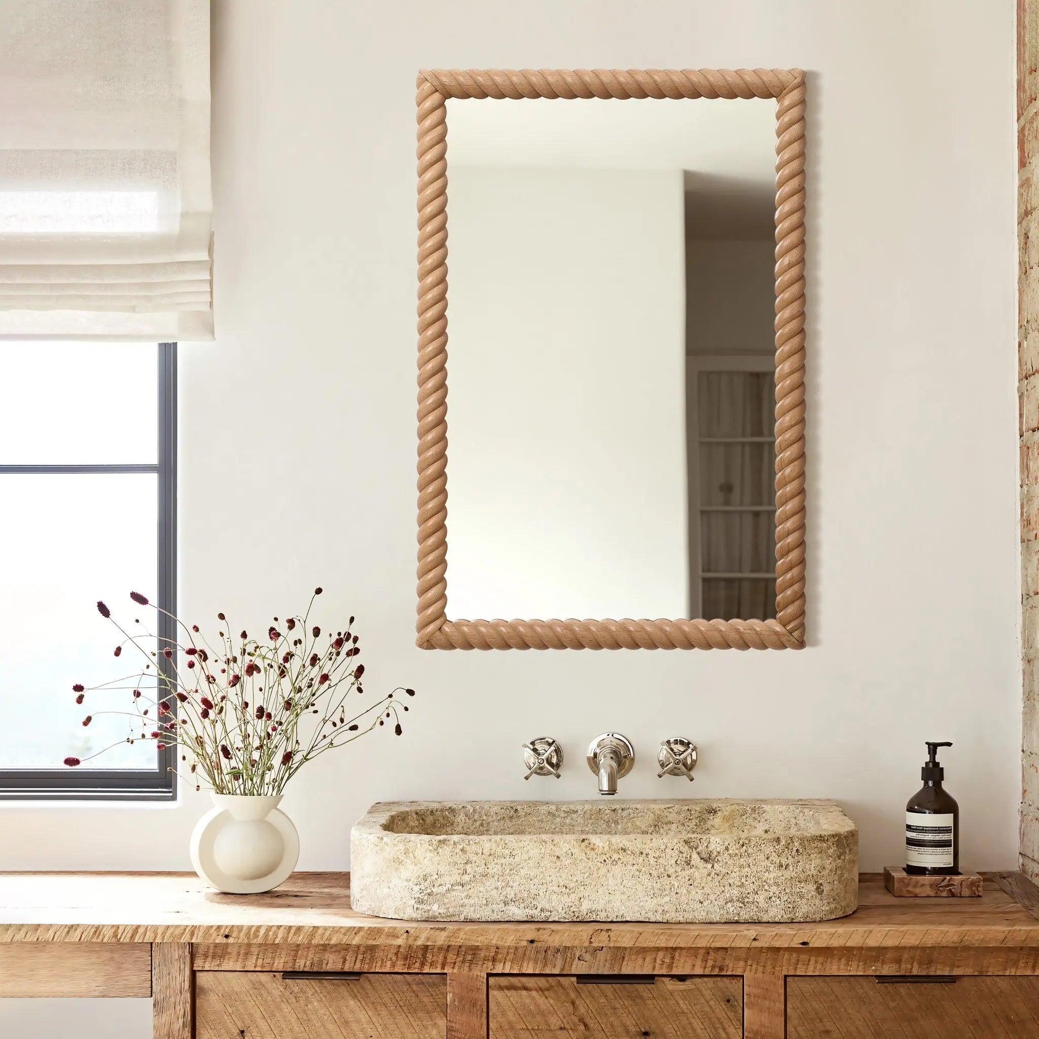 Bathroom with a rustic wooden vanity, stone sink, and elegant decorative wall mirror. A vase with flowers and soap dispenser sit on the counter. A window with sheer curtains is partially visible, adding a touch of elegance to the home decor.