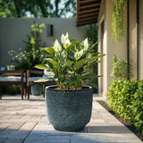 A LuxenHome Marina gray glazed round ceramic planter, 13.98 h, with lush green leaves sits on a sunny patio. The backdrop includes a wooden dining set and greenery, highlighted by the planters gradient color, creating a peaceful outdoor scene with natural elements.