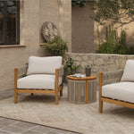 Outdoor patio scene with two Outdoor Acacia Wood Armchairs featuring weather-resistant cushions, a round table with decor, a textured rug, and a stone wall with greenery in the background.