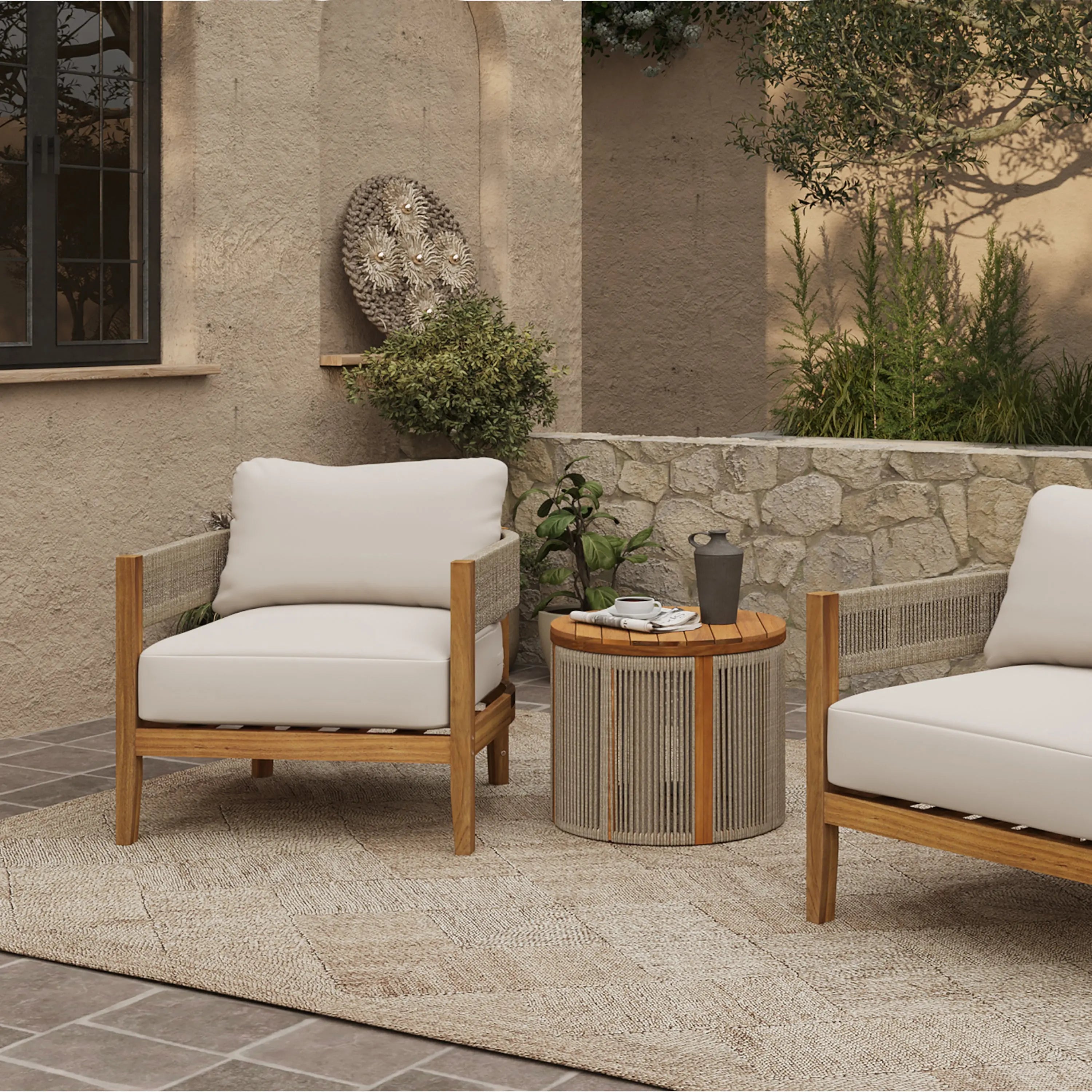 Outdoor patio scene with two Outdoor Acacia Wood Armchairs featuring weather-resistant cushions, a round table with decor, a textured rug, and a stone wall with greenery in the background.