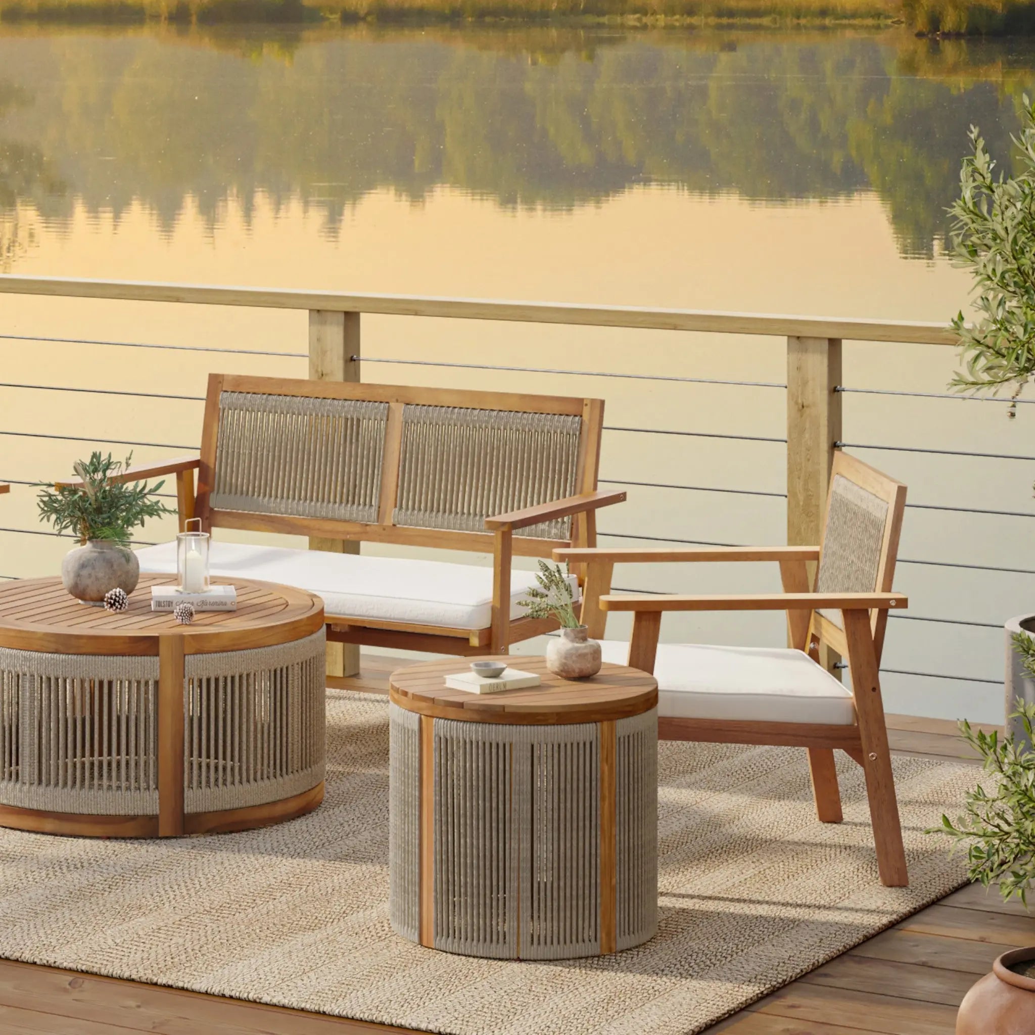 An outdoor patio featuring wooden furniture, including a loveseat and chair crafted from natural acacia wood, alongside two drum-shaped round end tables. The set rests on a hand-woven rope rug near a railing that overlooks the calm lake.