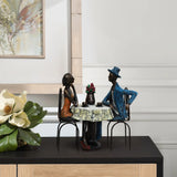A modern brown resin sculpture captures a romantic moment of two seated figures, one in a blue hat and suit, the other in an orange dress, at a table. Ideal as a wedding gift, it features an elegant flower arrangement on a black surface.
