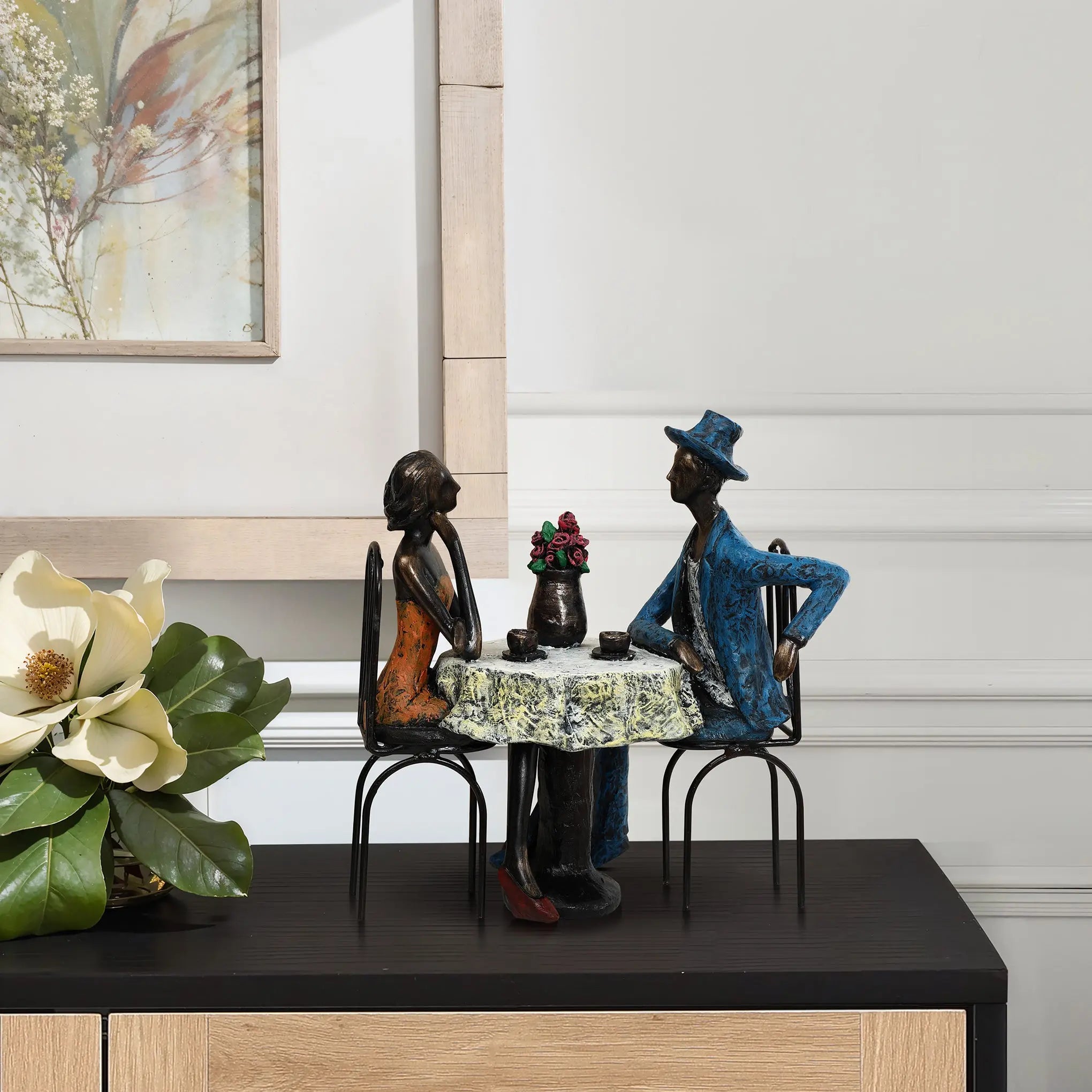 A modern brown resin sculpture captures a romantic moment of two seated figures, one in a blue hat and suit, the other in an orange dress, at a table. Ideal as a wedding gift, it features an elegant flower arrangement on a black surface.