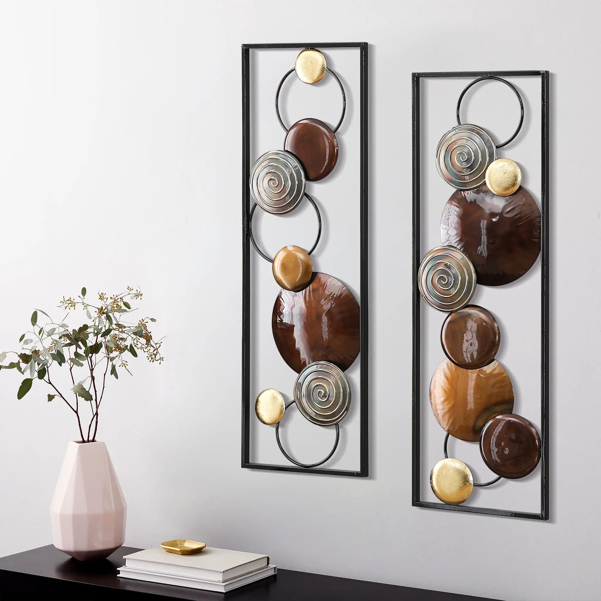 Two rectangular handcrafted panels feature circular and spiral designs in brown, gold, and silver tones. This metal wall decor hangs above a black console table with a pink vase and stacked books, perfect for contemporary home décor.