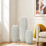 In a well-lit living room, three LuxenHome Modern geometric pattern resin tall vases (13.5 high), with a braided antique effect, stand near a beige sofa adorned with yellow cushions. Above them, a framed picture of horses enhances the wall.