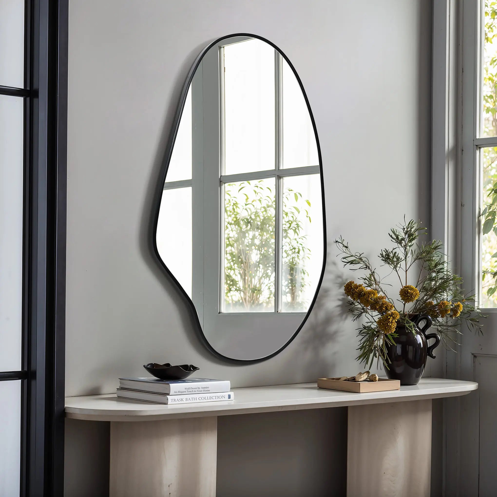 An irregular oval mirror with a black frame hangs above a console table, exuding a modern aesthetic. Books and a vase of yellow flowers rest below, creating an inviting vignette.