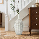 The LuxenHome Modern Geometric Pattern Resin Tall Trumpet Vase, standing 19.5 inches high, graces the wooden floor near a staircase with its elegant hand-painted design. Its tall narrow neck features branches, while a wooden cabinet with drawers sits to the right, creating a cozy and elegant decor.