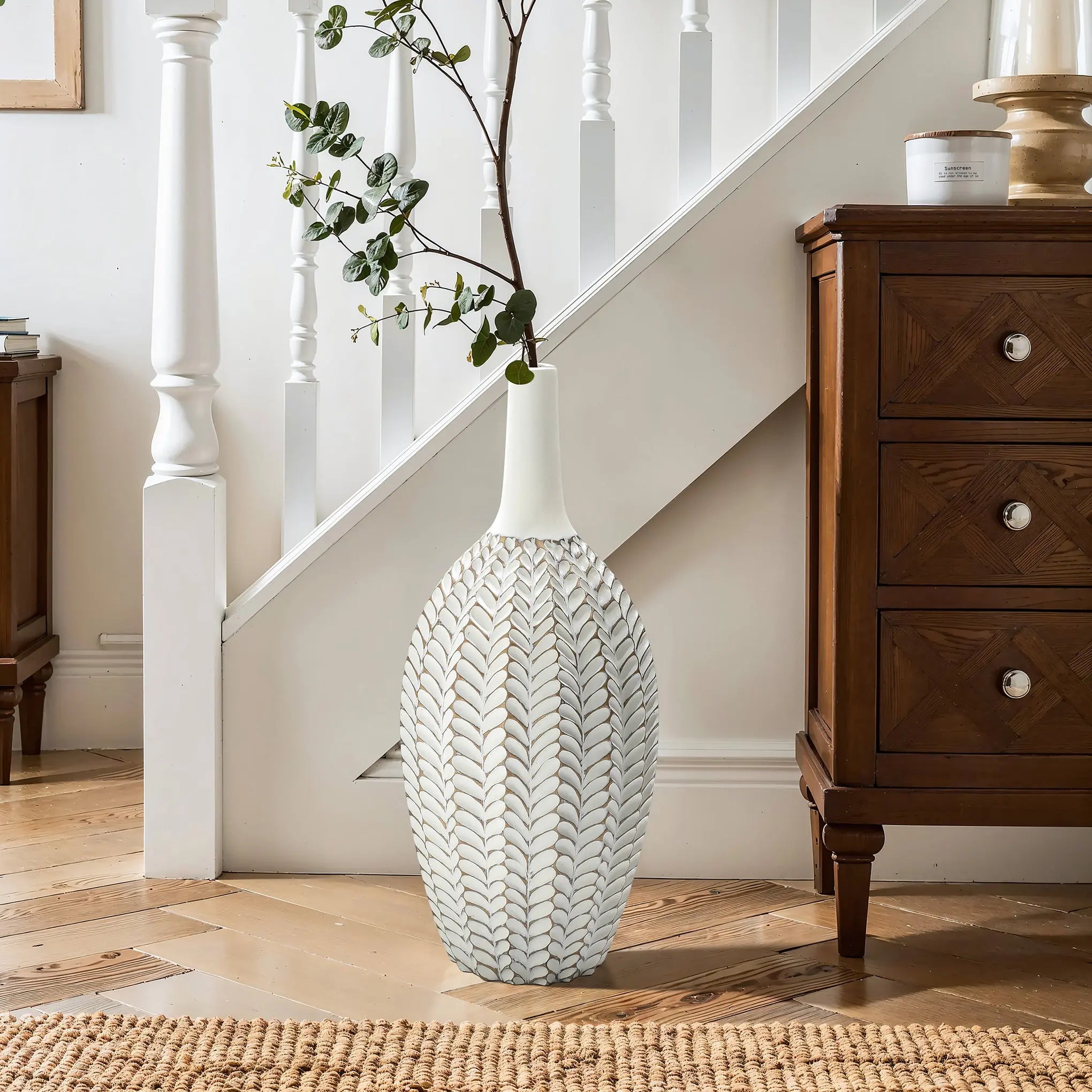 The LuxenHome Modern Geometric Pattern Resin Tall Trumpet Vase, standing 19.5 inches high, graces the wooden floor near a staircase with its elegant hand-painted design. Its tall narrow neck features branches, while a wooden cabinet with drawers sits to the right, creating a cozy and elegant decor.