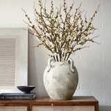 A LuxenHome rustic gray and white terracotta vase with handles, 11 high, holds branches of white blossoms on a wooden table. Nearby, a black bowl rests on Vintage Table Decoration. The background features a textured wall adorned with framed art for contemporary elegance.