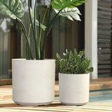 Two white, contemporary planters on a wooden deck: one with a tall plant, the other with a shorter plant, showcase their durable design near a pool and building.