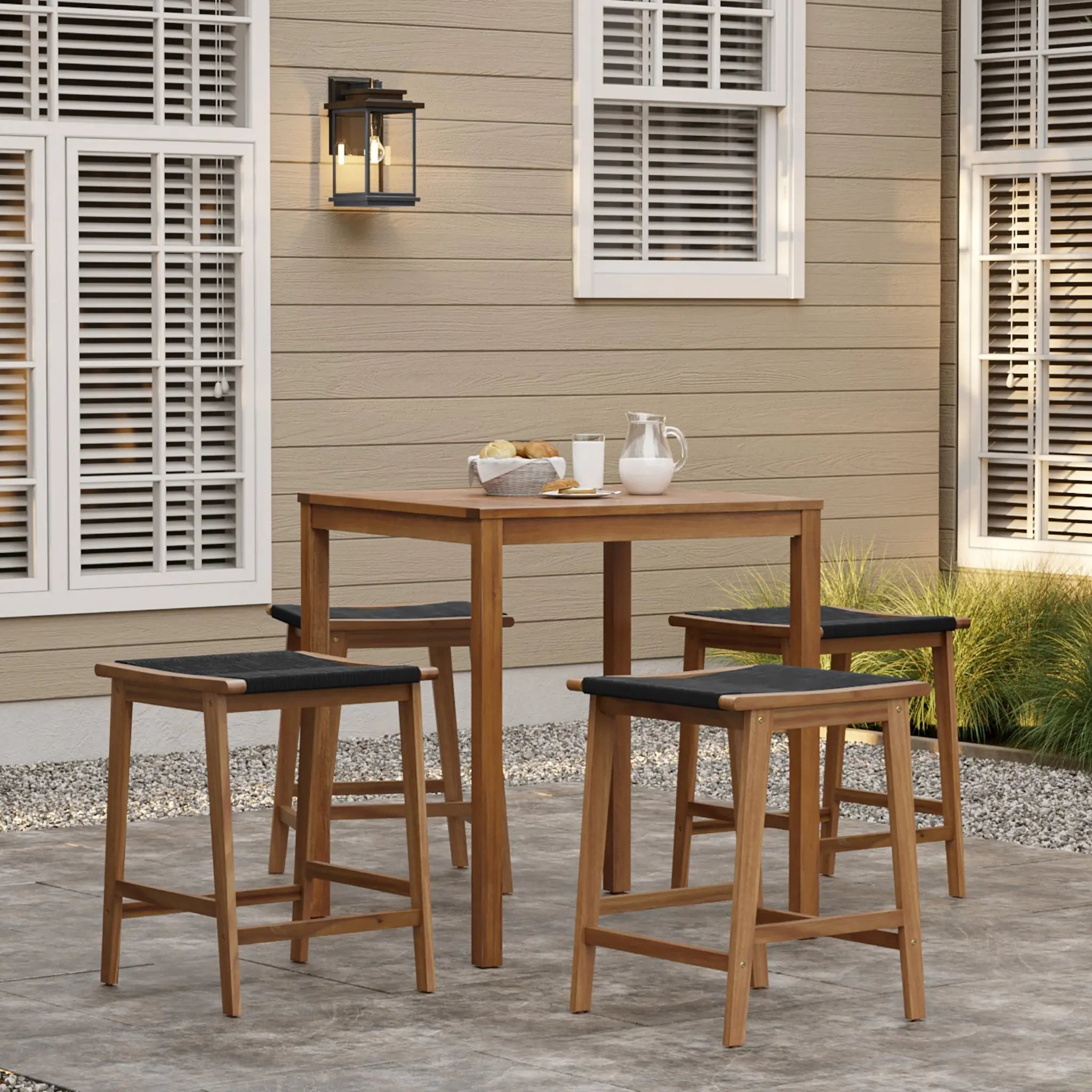 Outdoor patio scene with an FSC-certified acacia wood dining table and four matching stools on a stone surface. The table, perfect as an outdoor Acacia Wood Dining Table, holds a tray with food, a glass, and a pitcher. A tan building with white windows and a lantern completes the backdrop.