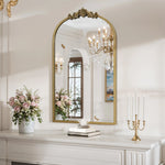 An elegant room showcases the LuxenHome Rêverie vintage gold accent metal frame arch mirror reflecting a chandelier and painting. A white mantel beneath holds a vase of pink flowers, candle holders, and stacked books, while soft lighting creates a warm ambiance.
