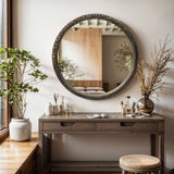A cozy room features a wooden desk adorned with vases, reed diffusers, and magazines. An Aged Wood Finish Crater Pattern Round Wall Mirror by LuxenHome reflects part of the room. A large potted plant by the window adds greenery to this serene space with its subtle industrial-inspired design.
