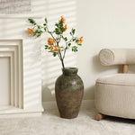 A textured stoneware vase with orange flowers on a branch sits on a light-colored floor beside a beige chair and a white fireplace. Shadows from blinds cast patterns, enhancing the rustic charm of this peaceful setting.