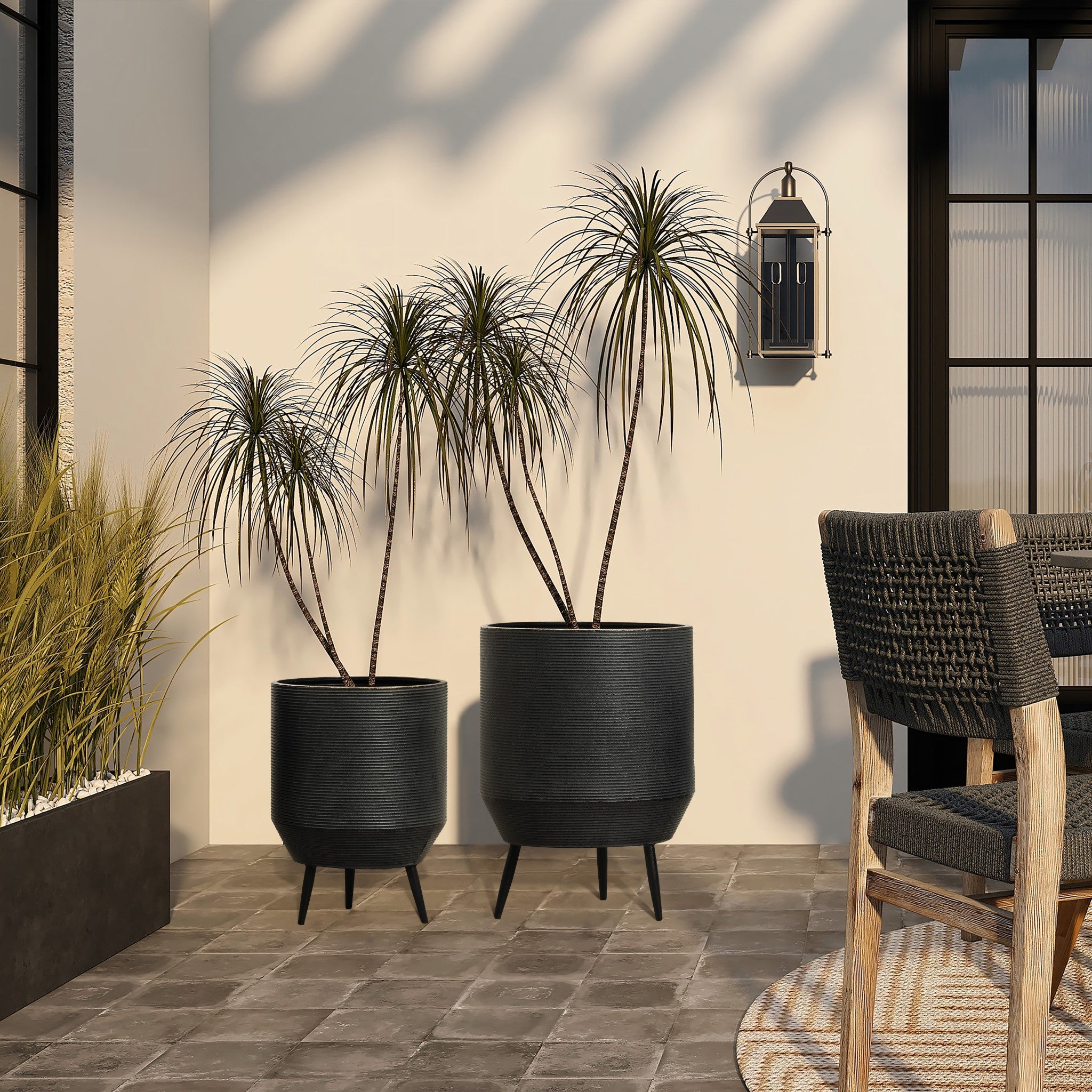 An outdoor scene showcases two potted plants in a pot planter set with a distressed black finish. A wicker chair and brown table add comfort, while a wall-mounted lantern casts a warm glow through large windows on the tiled patio.