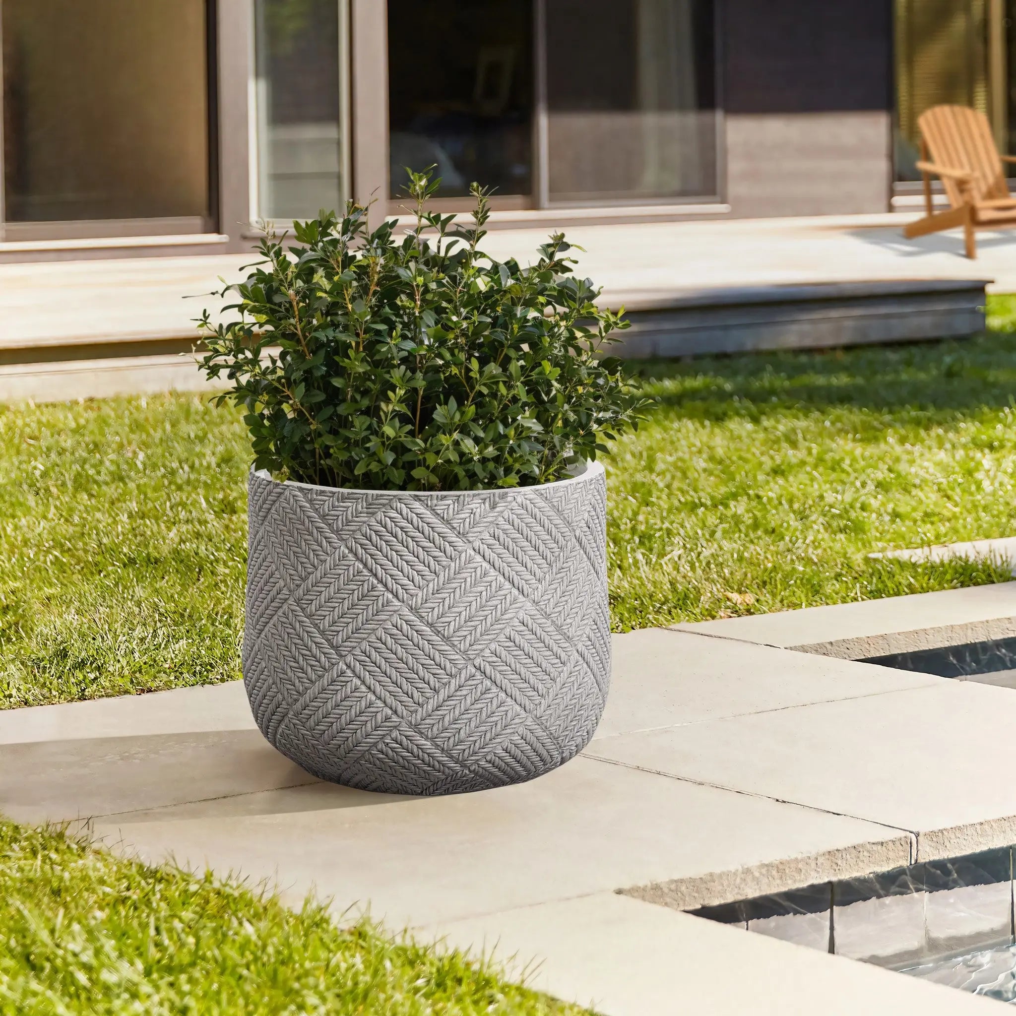 A green plant thrives in a textured gray pot crafted from durable MgO material on a patio. Nearby, a lawn and small water feature complement the scene, with modern building windows adding to the modern minimalist aesthetic.