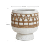 A round, textured white planter, crafted from magnesium oxide with Bohemian style brown wicker accents, measures 11.02 inches wide and 11.81 inches tall.